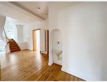 Heller Innenraum mit Holzdielenboden, weiß gestrichenen Wänden und Treppen aus Holz, die zu einem Fensterbereich führen. Rechts befindet sich ein kleines Waschbecken in einer Wandnische, und ein Heizkörper ist neben einer Türöffnung sichtbar.