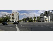 Straßenkreuzung in einer städtischen Umgebung mit mehrstöckigen Wohn- und Geschäftshäusern im Hintergrund, einem U-Bahn-Schild, mehreren geparkten und fahrenden Autos sowie grünen Bäumen entlang der Straße. Die Szene ist bei Tageslicht und unter einem leicht bewölkten Himmel aufgenommen.