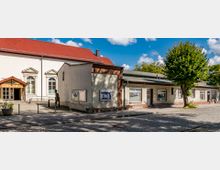 Außenansicht des Hans Claret Hauses, eines Gebäudes mit weißer Fassade, rotem Satteldach und klassizistischen Fensterelementen. Daneben befindet sich ein flacher Anbau mit beiger Fassade, Informationskästen und mehreren Fenstern, umgeben von einer gepflasterten Fläche und einem Baum.