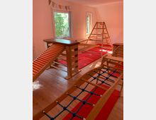 Ein heller Raum mit Holzboden, ausgestattet mit Spiel- und Klettergeräten aus Holz, wie einer Sprossenleiter, einer Rampe und einem Kletternetz mit blauen Seilen. Durch große Fenster fällt Tageslicht, und rote Matten liegen auf dem Boden.