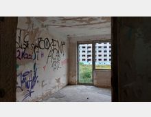Verlassener Raum mit grob verputzten Wänden, die mit Graffiti bedeckt sind. Durch ein großes, beschädigtes Fenster sind überwuchertes Gras und ein leerstehendes, graues Wohngebäude mit mehreren Fensteröffnungen sichtbar.
