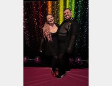 Indoor photo booth setup with a rainbow-striped, star-speckled backdrop and a pink floor, decorated with scattered balloons and tinsel along the base.