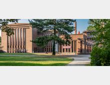 Backsteingebäude im Bauhaus-Stil mit großen vertikalen Fenstern und einem hohen Schornstein, umgeben von Bäumen und einer Rasenfläche. Ein Banner mit der Aufschrift „Demokratie“ hängt über dem Eingang, und ein Weg führt durch einen parkähnlichen Bereich zum Gebäude.