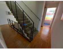 Ein Innenraum mit einer Treppe aus Beton und Metallgeländern, die neben einem Flur mit Holzboden verläuft. Im Hintergrund ist ein Badezimmer mit einem Duschvorhang in bunten Streifen sichtbar.