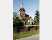 Historische Dorfkirche mit Fachwerk-Turm und rotem Ziegeldach, umgeben von Bäumen und einer niedrigen Ziegelsteinmauer, im Vordergrund ein steinernes Kreuz.