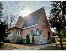 Ein Einfamilienhaus mit rosafarbener Fassade und grauem Ziegeldach, umgeben von Bäumen und einem gepflasterten Weg. Das Gebäude besitzt einen gemauerten Sockel aus Naturstein und eine verglaste Veranda.