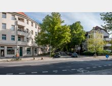 Straßenecke in einem Wohngebiet mit einem modernen mehrstöckigen Gebäude links, Geschäften im Erdgeschoss und einem Baumgesäumten Straßenabschnitt im Hintergrund, daneben ein weiteres Wohngebäude mit Balkonen. Fahrräder und geparkte Autos sind sichtbar, sowie Verkehrsschilder und Straßenmarkierungen.