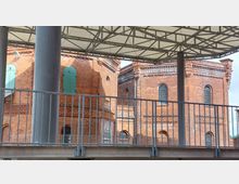 Backsteingebäude mit historischen Details wie grünen Fensterläden und verzierten Mauerwerken, teilweise durch eine Metallbrüstung und eine moderne Überdachung mit Metallträgern verdeckt. Die Szene zeigt einen Mischstil aus Industriearchitektur und moderner Konstruktion.