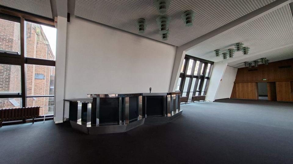 Großer, leerer Innenraum mit dunklem Boden, einer zentralen Theke aus Metall und großen Fensterfronten, die den Blick auf ein Backsteingebäude freigeben. Die Decke ist mit rasterförmigen Paneelen und hängenden Leuchten ausgestattet, am Ende des Raums befindet sich eine holzverkleidete Wand mit Durchgang.