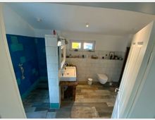 Modernes Badezimmer mit weiß gefliesten Wänden, einer bodengleichen Dusche mit türkis-blauen Fliesen auf der linken Seite, einem Waschtisch mit einem rechteckigen Waschbecken und einem hölzernen Unterschrank in der Mitte. Rechts befindet sich eine hängende Toilette, und ein kleines Fenster lässt Tageslicht in den Raum. Der Boden ist mit grauen und braunen Fliesen gestaltet.