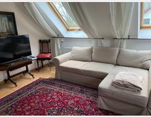 Wohnzimmer mit einer beigen Eckcouch, einem roten Orientteppich, einem Holztisch mit einem Fernseher darauf und einem antiken Stuhl mit rotem Sitzpolster. Im Hintergrund Dachfenster mit weißen Vorhängen und Blick ins Freie.