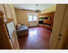 Schmales Wohnzimmer mit gestreifter Tapete, rotem Dielenboden und Deckenleuchte; an der Stirnseite ein Fenster mit Heizkörper darunter. Rechts steht eine große dunkle Wohnwand mit Fernseher, links ein Sofa, im Vordergrund ist der Eingang mit Kachelofenansicht zu sehen.