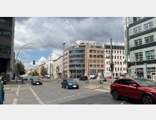Straßenszene in einer Stadt mit mehrspuriger Kreuzung, moderner und historischer Bebauung. Im Vordergrund fahren Autos, während Fußgänger an Ampeln warten; im Hintergrund ist ein rundes Glasgebäude sichtbar, flankiert von älteren Wohnhäusern.