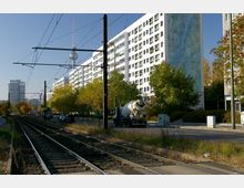 Straßenszene in einer Stadt mit Straßenbahngleisen im Vordergrund, flankiert von mehrstöckigen Wohngebäuden auf der rechten Seite. Im Hintergrund ist der Berliner Fernsehturm sichtbar, umgeben von Herbstbäumen und weiteren Hochhäusern.