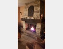 Ein gemauerter Kamin in einem rustikalen Raum, mit brennendem Feuer und Holzscheiten. Auf dem breiten Kaminsims stehen Kerzenhalter und Dekorationen, darunter eine kleine Weihnachtsfigur.