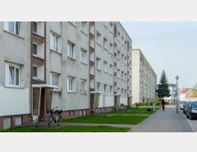 Mehrstöckiges Wohngebäude aus Beton mit einfachen Fassaden, mehreren rechteckigen Fenstern und kleinen Eingangsbereichen mit Überdachungen. Vor dem Gebäude befinden sich ein Grünstreifen, Gehwege, Fahrradständer und parkende Autos; im Hintergrund sind weitere Häuser und ein Baum zu sehen.