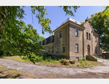 Alt-Text: Zweistöckiges historisches Gebäude mit einer Putzfassade und einem Giebel über dem Eingang, umgeben von Bäumen. Ein Weg führt an der Vorderseite entlang, teilweise von Grünflächen und Baumstümpfen gesäumt.