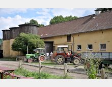 Bauernhof mit gelbem Gebäude und roten Ziegeldach, zwei Traktoren im Vordergrund und mehreren Holzsilos. Ein Baum steht neben dem Gebäude, und Personen sind in der Nähe eines Eingangstors zu sehen.