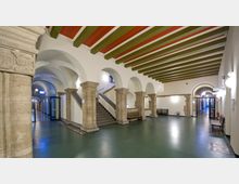 Ein historischer Innenbereich mit Rundbögen, massiven Steinsäulen und einer grünen Decke mit roten und grünen Streben. Eine Treppe führt nach oben, und der Boden besteht aus dunkelgrünem Material, während die Gänge von Wandleuchten beleuchtet werden.