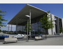 Modernes Gebäude mit großen Glasfassaden und einem auskragenden Flachdach, umgeben von Gehweg, Bäumen und geparkten Autos. Im Vordergrund sind Sitzbänke und Laternen zu sehen.