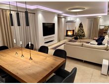 Modernes Wohnzimmer mit Essbereich, einem hölzernen Esstisch mit Kerzenhaltern und schwarzen Stühlen. Im Hintergrund ein Sofa, ein Weihnachtsbaum, ein brennender Kamin und ein Fernseher an der Wand; helle Vorhänge und dezente indirekte Beleuchtung prägen den Raum.