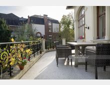 Eine Terrasse mit einem Tisch und geflochtenen Stühlen, umgeben von einem metallenen Geländer mit Pflanzen in Töpfen. Im Hintergrund sind Bäume und ein historisches Backsteingebäude sichtbar.