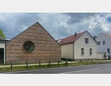 Straßenansicht mit einem modernen Gebäude aus Klinkersteinen mit einem großen runden Fenster auf der Fassade und daneben zwei einfache Wohnhäuser mit roten Ziegeldächern und weißer Putzfassade. Im Vordergrund ein Gehweg mit Pollern und eine Straße, im Hintergrund bewölkter Himmel.