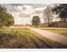 Weitläufiges, parkähnliches Gelände mit einem Kiesweg und Schienen, umgeben von Bäumen und Wiesen. Im Hintergrund sind ziegelrote Gebäude und eine alte Backsteinmauer zu sehen, unter einem bewölkten Himmel.