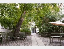 Ein gepflasterter Hof mit grünen Metallstühlen und -tischen, eingerahmt von dichten Bäumen und Sträuchern. Ein breiter Gehweg führt durch die Mitte, und im Hintergrund ist eine Türöffnung in einem weißen Gebäude sichtbar.