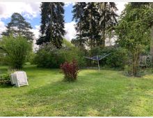 Weitläufiger Garten mit Rasenfläche, großen Bäumen und blühenden Sträuchern. Im Vordergrund stehen weiße Plastikstühle gestapelt, daneben ein Wäschespinnengerüst und ein gerollter Gartenschlauch an einem Zaun im Hintergrund.