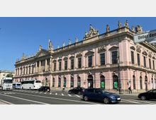 Historisches Gebäude mit einer rosa Fassade und ornamentaler Architektur, gekrönt von mehreren Statuen. Im Vordergrund fahren Autos und Busse auf einer belebten Straße, daneben befindet sich ein Fußgängerbereich.