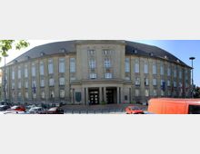 Großes, historisches Gebäude mit steinerner Fassade, rechteckigen Fenstern und einem geschwungenen Dach. Der Eingangsbereich ist mit Säulen und großen Türen gestaltet, davor parken mehrere Autos, und es sind Flaggen aufgestellt.