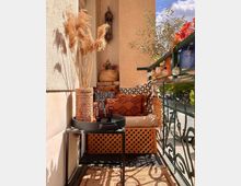 Kleiner Balkon mit gemütlicher Sitzecke, dekoriert mit Kissen in warmen Farben, Trockenpflanzen in einer Vase, einer orientalischen Laterne und einer Tischablage mit Gläsern. An der Metallbrüstung hängen kleine Lichterketten, daneben Pflanzen und Herbstdekoration.