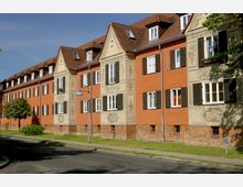 Mehrstöckiges Wohngebäude mit roter Fassade und verzierten Sandsteinelementen, das schräg entlang einer ruhigen Straße mit Grasstreifen verläuft. Die Fenster sind von braunen Fensterläden eingerahmt, und das Dach ist mit roten Ziegeln gedeckt.