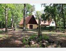 Ein gelbes Landhaus mit roten Ziegeldächern und einem Anbau, umgeben von hohen Bäumen in einem bewaldeten Areal. Im Vordergrund liegen gefällte Baumstämme, und ein schattiger Gartenbereich mit Tischen und Stühlen ist sichtbar.