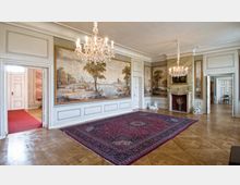 Historischer Innenraum eines prunkvollen Raumes mit Holzboden, verzierten Wandmalereien, einem roten Orientteppich und zwei funkelnden Kronleuchtern. Die Wände sind mit Wandpaneelen und einem dekorativen Kamin gestaltet, flankiert von Statuen, durch Türen sind angrenzende Räume sichtbar.