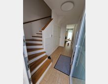 Ein moderner Flur mit hellem Fliesenboden, einer offenen Holztreppe mit weißem Geländer und einer Glastür. Im Hintergrund sind ein Wohnbereich mit Tageslicht und Dekorationselementen sichtbar.