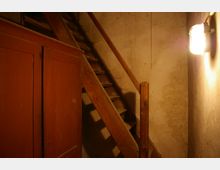 Ein einfacher Raum mit rohem Betonputz an den Wänden, einer Holztreppe mit Geländer und einem roten Holzschrank im Vordergrund. Eine Wandlampe mit Käfigschutz beleuchtet den Bereich.