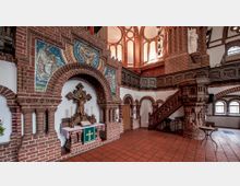Innenansicht einer Backsteinkirche mit reich verzierten Bögen und Wandmosaiken. Zu sehen sind ein Altar mit Kruzifix, seitliche Holzkanzeln, Arkaden und helle Fenster, die den Raum erleuchten.