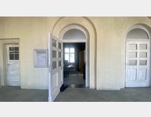 Ein Korridor mit hellen Wänden und drei Türen, von denen die mittlere geöffnet ist und in einen Raum mit einem Fenster und Möbeln führt. Links neben der mittleren Tür befindet sich ein Schild mit Wegweisern zu verschiedenen Büros.