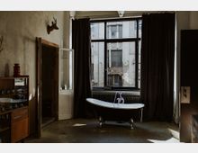 Ein industriell gestalteter Innenraum mit einer freistehenden Badewanne vor einem großen Fenster, das eine Backsteinfassade zeigt. Der Raum hat dunkle Vorhänge, eine Wand mit einem Hirschkopf-Dekor und eine Vitrine aus Holz auf der linken Seite.