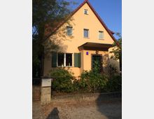 Ein zweigeschossiges Haus mit gelb verputzter Fassade, rotem Satteldach und grünen Fensterläden, umgeben von einem Garten mit niedriger Hecke und Bäumen im Hintergrund. Vor dem Haus befindet sich ein kleiner Kiesweg mit einem Briefkasten an der linken Seite.