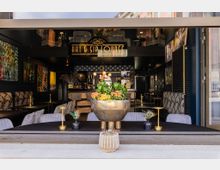 Modernes Café oder Lounge mit offener Glasfront, dekoriert mit kunstvollen Gemälden an den Wänden und gemütlichen Sitzbereichen. Ein großer goldener Pflanzenkübel steht im Vordergrund auf einem Tisch, umgeben von kleinen dekorativen Vasen und Kerzenhaltern.