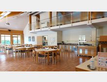 Ein Speisesaal mit Holzmöbeln, runden und rechteckigen Tischen, sowie einem großen Buffetbereich mit Glasschutz und Metallfronten. Im Hintergrund sind Fenster mit bunten Vorhängen und eine Empore mit Glasgeländer zu sehen.