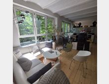 Ein modernes Büro mit hellem Holzfußboden und großen Fenstern, die Ausblick auf grüne Bäume bieten. Im Vordergrund befindet sich eine weiße Eckcouch mit runden Tischen, während der hintere Bereich Schreibtische, Stühle und Regale zeigt.