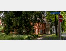 Straßenszene mit einem roten Gebäude mit grünen Fensterläden, umgeben von großen Bäumen und einer Grünfläche mit Blumen und Bänken. Ein Straßenschild mit der Aufschrift "Hevellerstraße" und ein Durchfahrtsverbotszeichen sind im Vordergrund sichtbar.