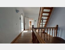 Hell erleuchtetes Treppenhaus mit Holzboden und einer Holztreppe auf der rechten Seite, die nach oben führt. Im Hintergrund ist ein geöffneter Flur mit mehreren Türen und einem Raum mit Holzmöbeln sichtbar.