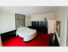 Ein Schlafzimmer mit rotem Teppichboden, einem runden Bett mit weiß gepolsterter Umrandung und einer großen schwarz-weißen Wanddekoration über dem Bett. Im Raum stehen schwarze Schränke und ein weißer Türrahmen mit einer geschlossenen Tür, daneben eine mittig platzierte Zimmerpflanze.