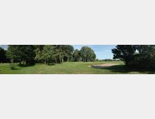 Weitläufige grüne Landschaft mit einer Rasenfläche, umgeben von Bäumen, einem Sandbunker auf der rechten Seite und einem leicht bewaldeten Bereich im Hintergrund. Der Himmel ist klar und blau.