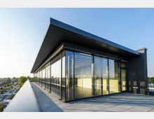 Modernes Gebäude mit Glasfassade und Flachdach auf einer großen Terrasse, die in eine urbane Umgebung mit Bäumen und Straßen eingebettet ist. Die Glasfronten reflektieren das Tageslicht, während die klare Architektur klare Linien und viel Transparenz zeigt.